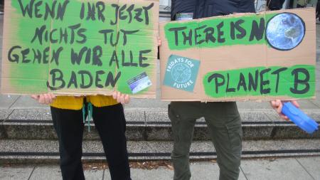 Two people with cardboard signs that read "There is no Planet B" and "wenn ihr jetzt nichts tut gehen wir alle baden"